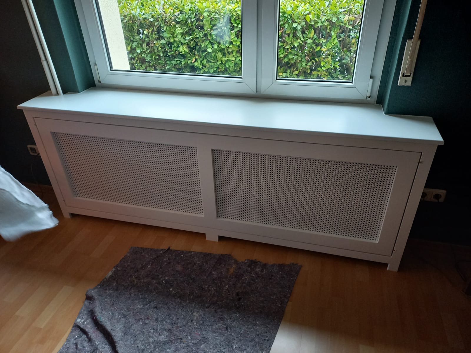 Weißer Heizkörperverkleidung vor einem Fenster mit Gittermuster auf hellem Holzboden