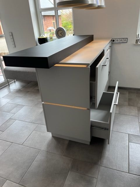 Eine moderne Kücheninsel in hellgrauer Farbe mit geöffneten Schubladen und LED Beleuchtung