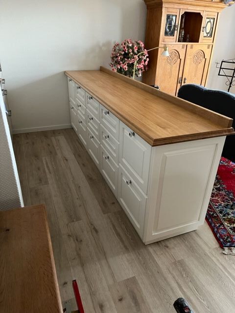 Eine lange weisse Kommode mit Holzplatte steht auf hellem Boden neben einem Teppich