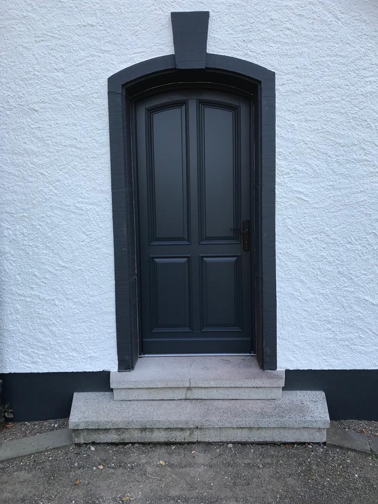 Eine dunkelgraue Tür mit vier Feldern in weißer Putzfassade und Steinstufen