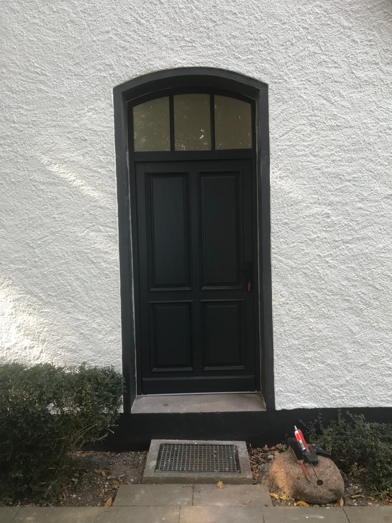 Eine schwarze Holztür mit Fenster ist in einer weißen verputzten Wand eingelassen