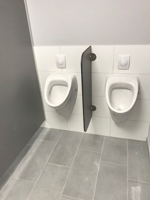 Zwei weiße Urinale mit grauer Trennwand stehen nebeneinander an einer gefliesten Wand