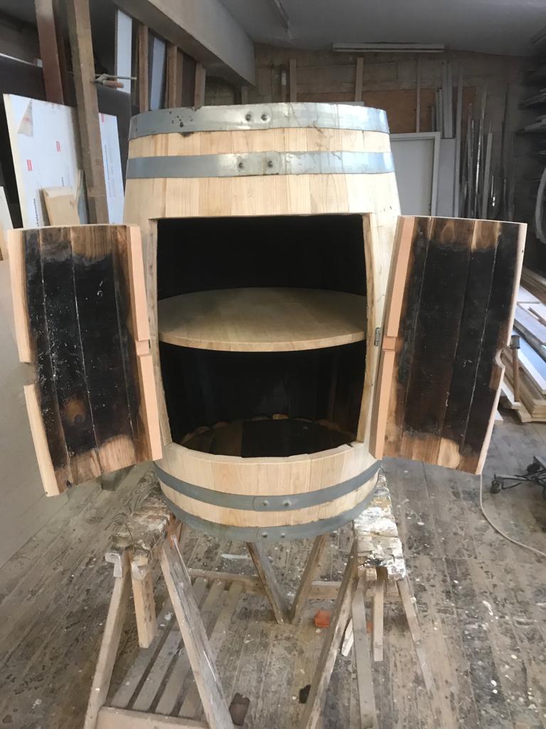 Ein Holzfass mit zwei geöffneten Türen, innen ein Regal und metallenen Fassringen